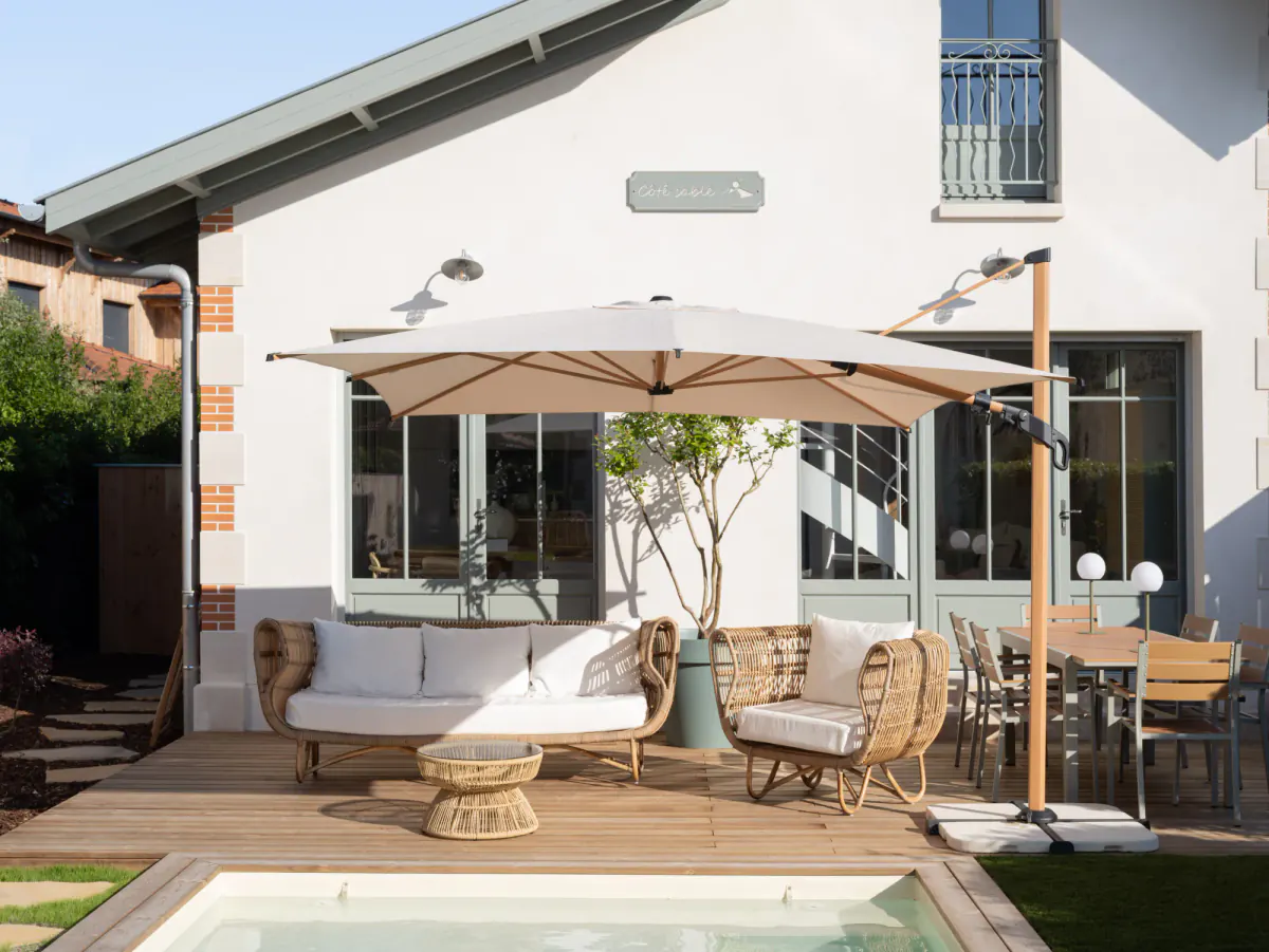 Terrasse de Côté Sable : canapé en rotin, parasol déporté et table de repas au bord de la piscine